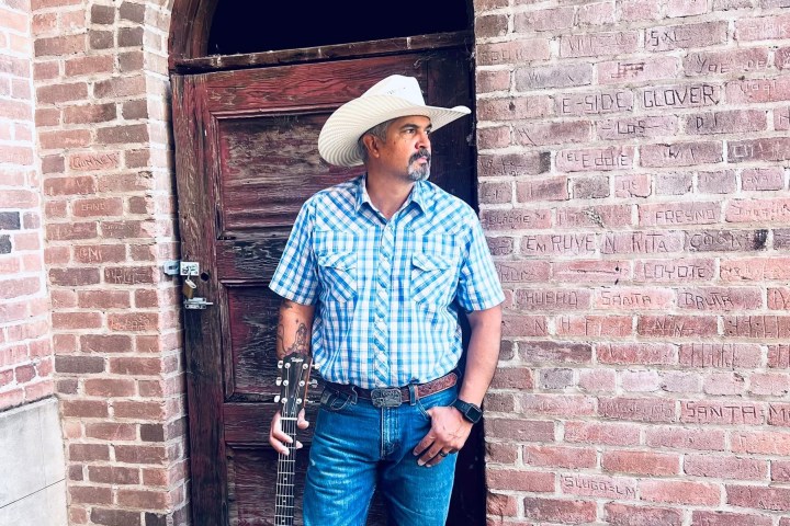 Man in cowboy hat holding guitar, standing by arched brick doorway.